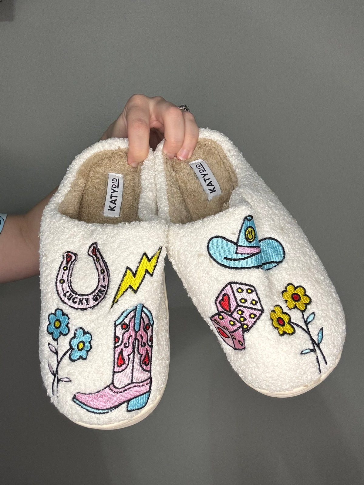 Pair of white slippers with colorful  cowgirl designs held by a hand against a gray background