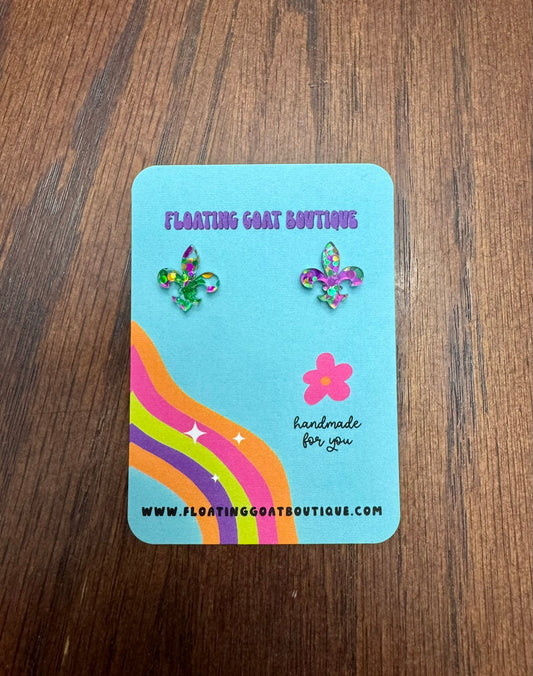 Pair of Fleur de Lis Mardi Gras Confetti earrings on a blue card with rainbow design on a wooden surface.