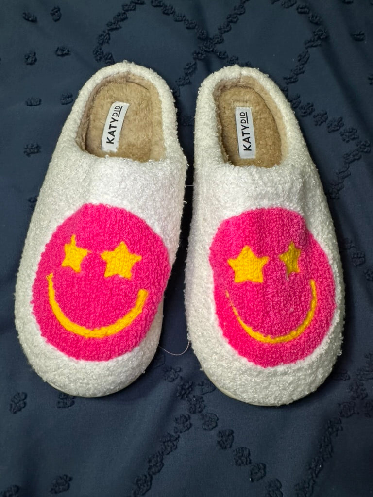 Pair of white slippers with pink smiley face with star eyes design on a dark fabric background