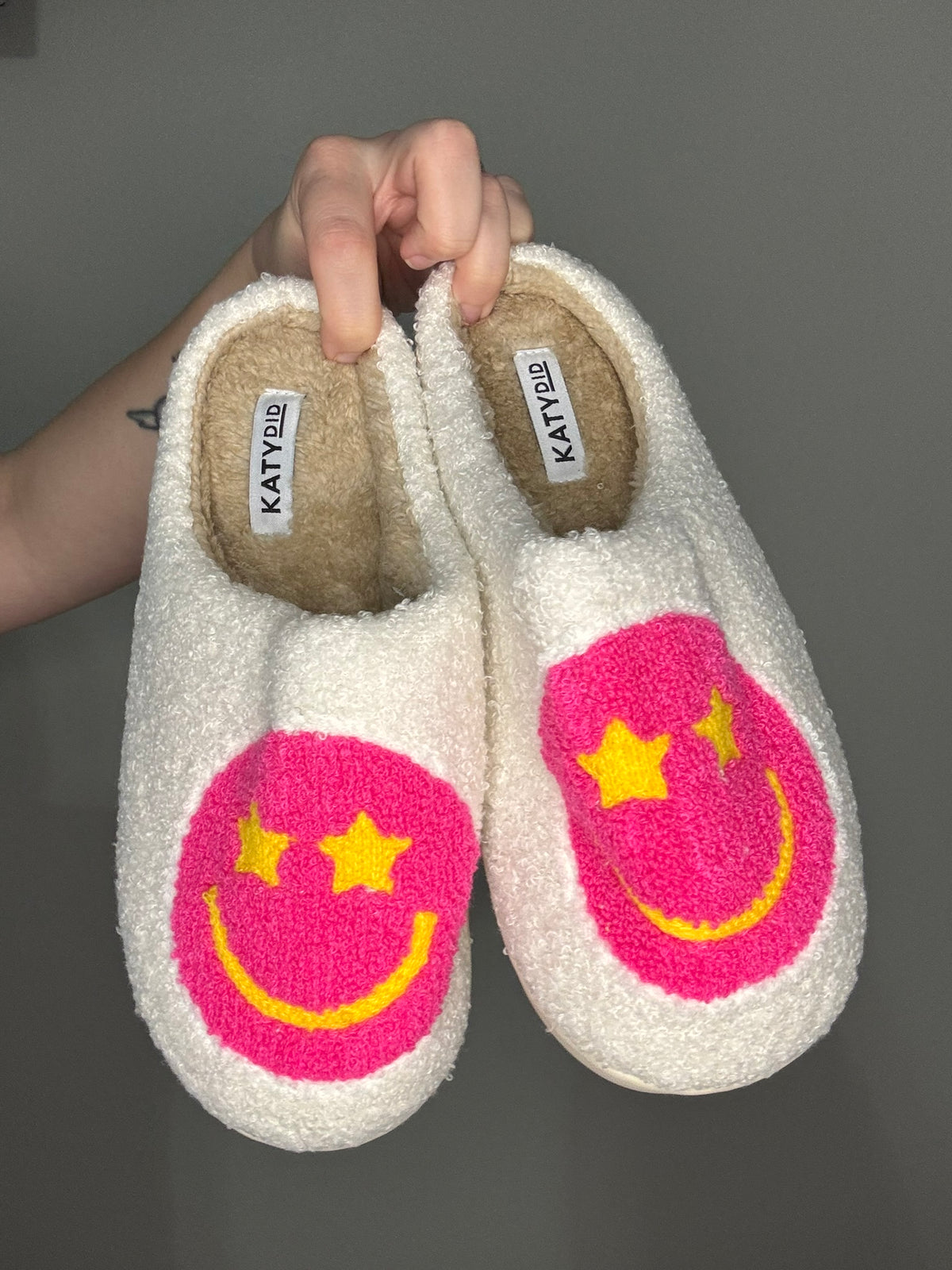 Pair of white slippers with pink smiley face with star eyes design held by a hand against a gray background